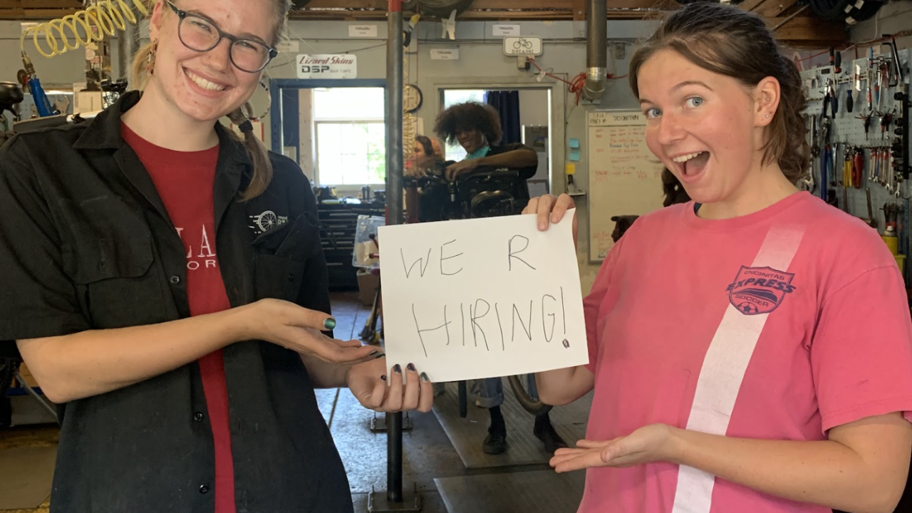 Two Bike Barn mechanics hold a sign that says "We R Hiring"