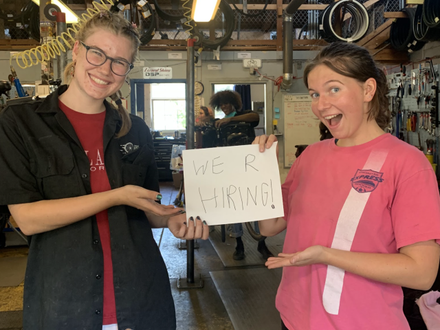 Two Bike Barn mechanics hold a sign that says "We R Hiring"